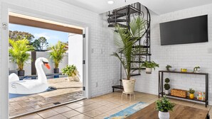 A modern living room with a white brick wall, a spiral staircase, a large TV, and a pool with a swan float. - Pool Oasis -Near RJ Stadium, Hyde Park & Downtown (Tampa)
