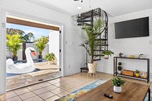 A modern living room with a white brick wall, a spiral staircase, a large TV, and a pool with a swan float. - Pool Oasis -Near RJ Stadium, Hyde Park & Downtown (Tampa)