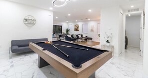 Una mesa de billar en una sala de estar moderna, con un sofá y un comedor al fondo.