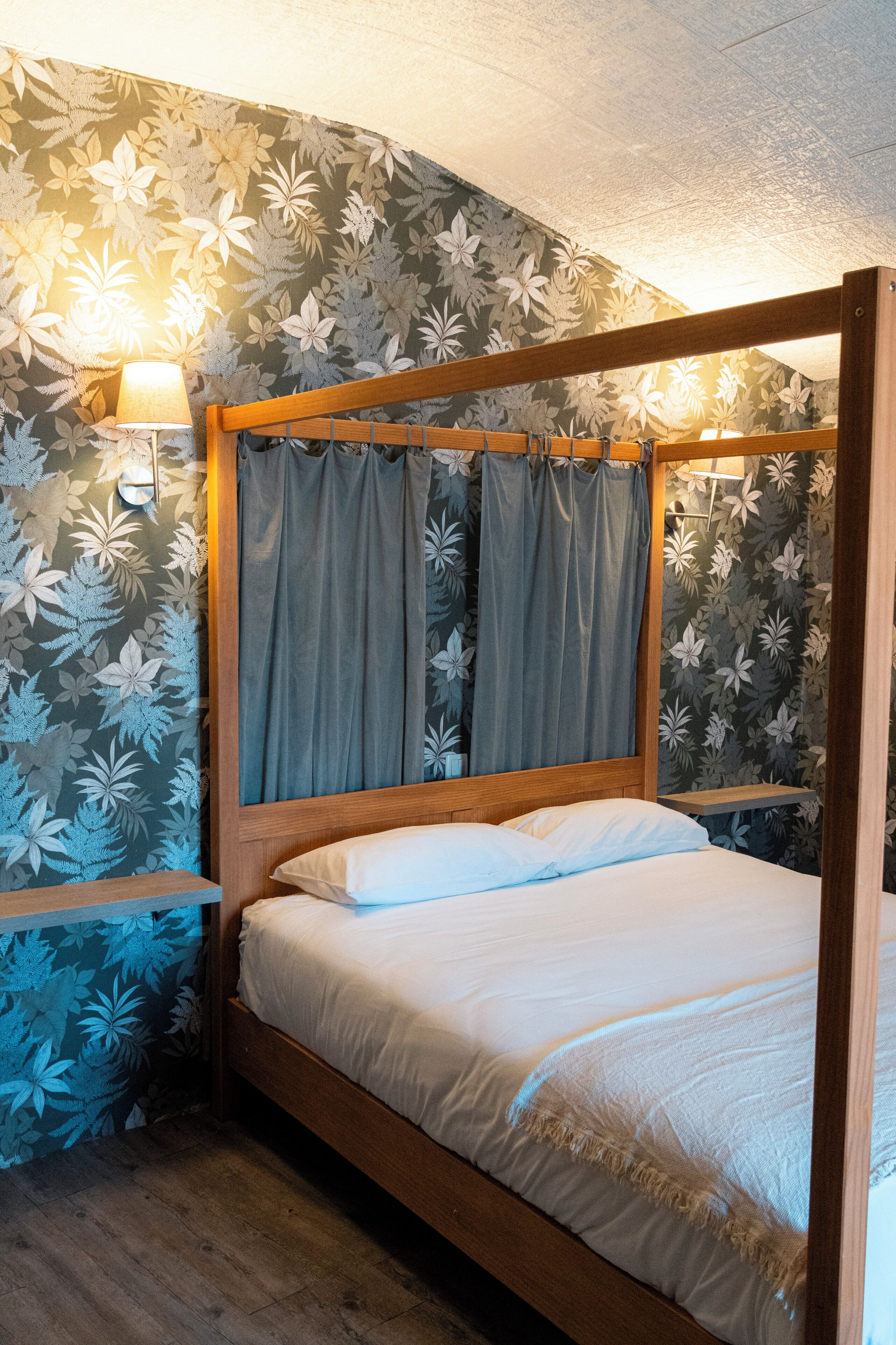 A four-poster bed with a wooden frame, grey curtains, and white bedding in a room with floral wallpaper.