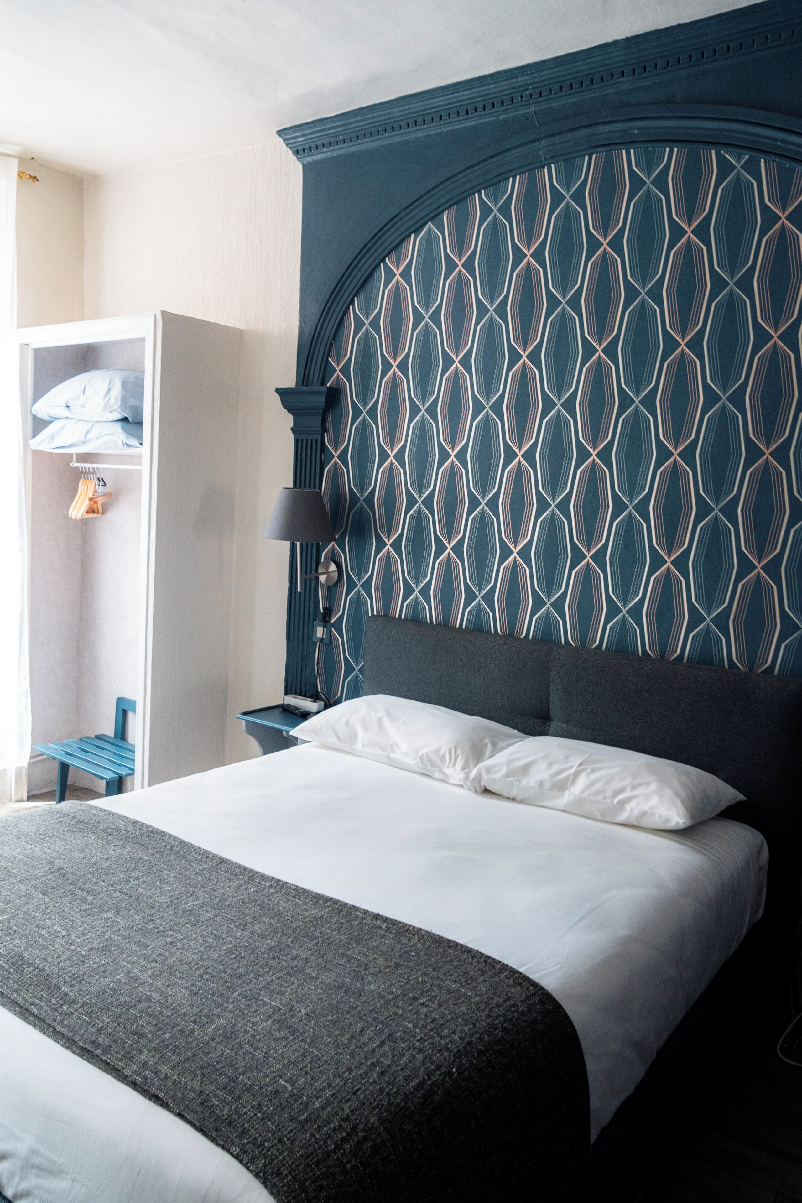 A neatly made bed with a dark headboard, a bedside table with a lamp, and a wardrobe with hanging clothes.