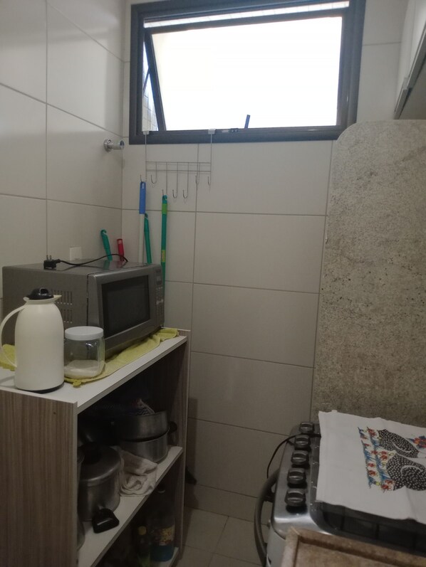 Uma cozinha compacta com micro-ondas, chaleira e utensílios sobre a bancada. Há uma janela acima da pia, e um fogão com um pano decorativo.