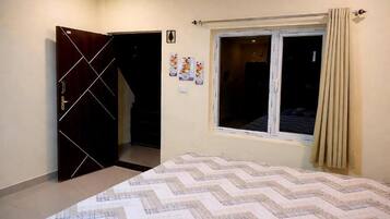 A bedroom with a bed, a window with curtains, a door, and a wall-mounted light switch.
