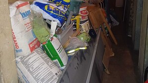 Een smalle gang met leidingen en een boiler aan de muur. Tassen met verschillende goederen staan tegen de muur gestapeld.