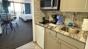 A compact hotel room with a kitchenette, a bed, a small dining area, and a view of the ocean.