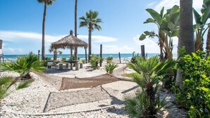 A beach area with a thatched gazebo, palm trees, and a pebble path leading to the sea.