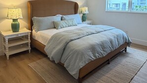 A bedroom with a bed, nightstands, a ceiling fan, a window with blinds, and a view of buildings outside. - Privateer Beachfront Retreat, Designer Renovation, Ground Floor Beachfront (Fort Myers Beach)