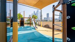 An outdoor playground with climbing structures and a slide, set against a city skyline. - Luxurious 1bedroom All Bills Included (Dubai)