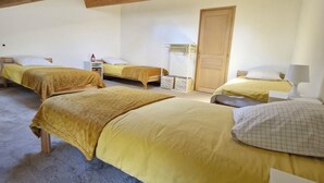 Una habitación con cuatro camas, vigas de madera en el techo, una puerta blanca y una pequeña mesa blanca con una lámpara.
