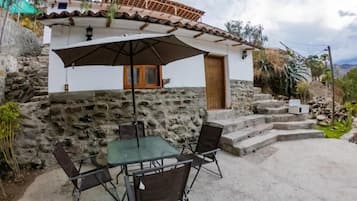 A stone house with a tiled roof, a wooden door, and a small window. There is a table with four chairs and a large umbrella outside.