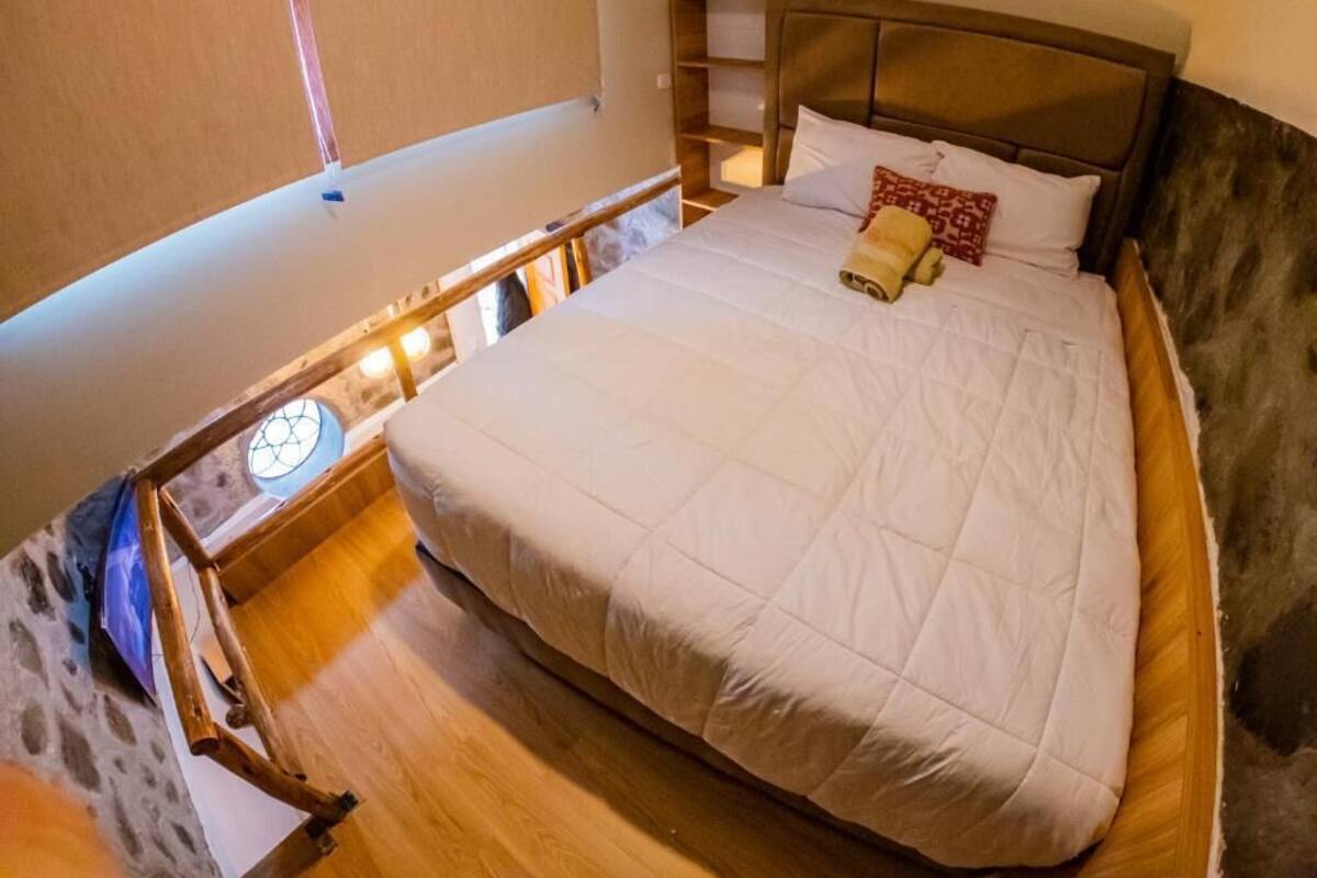 Een slaapkamer met een groot bed, een houten hoofdbord, een nachtkastje met een lamp en een stenen muur.