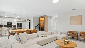 Un salon moderne avec un canapé d’angle, un coin repas avec une table et des chaises, et une cuisine en arrière-plan.