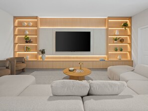 Ein modernes Wohnzimmer mit großem Flachbildfernseher, eingebauten Regalen mit Pflanzen, einem runden Couchtisch und einem beigefarbenen Sofa.