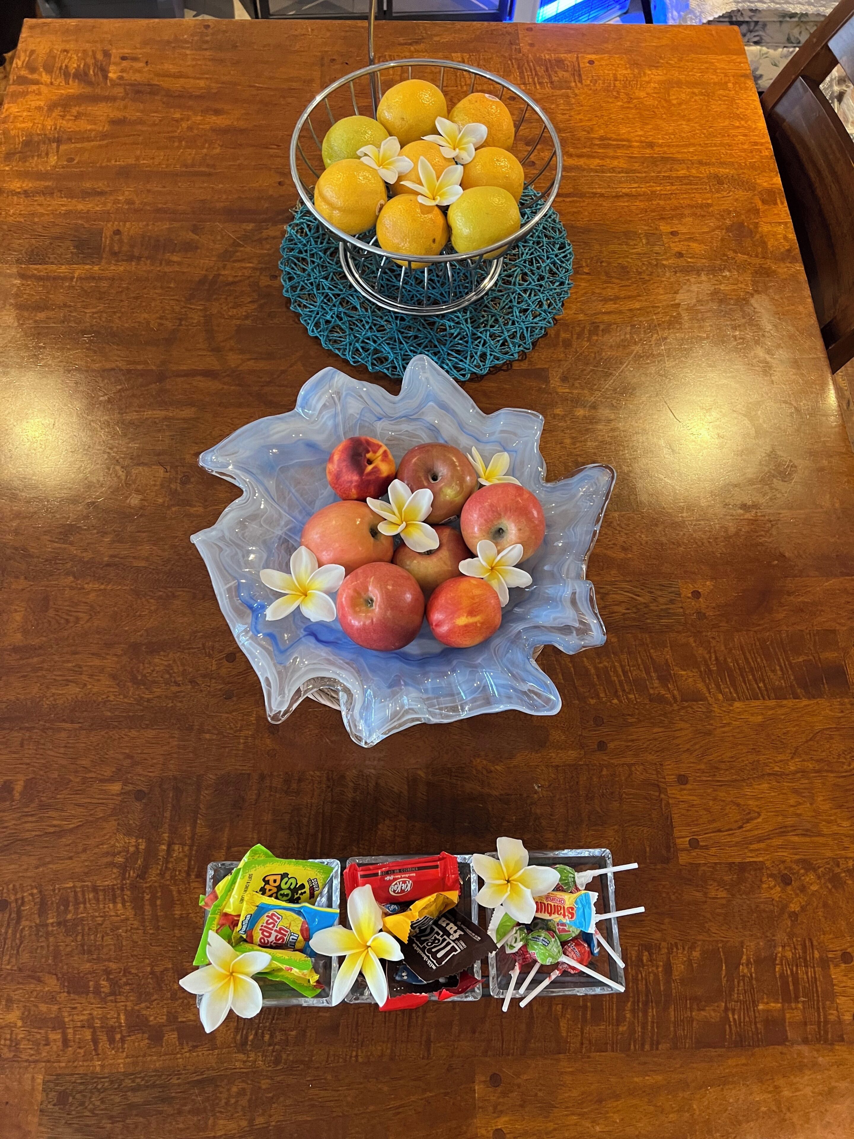 Een houten tafel met een schaal vol fruit, met sinaasappels en appels, een blauwe decoratieve schaal en een dienblad met snacks en bloemen.