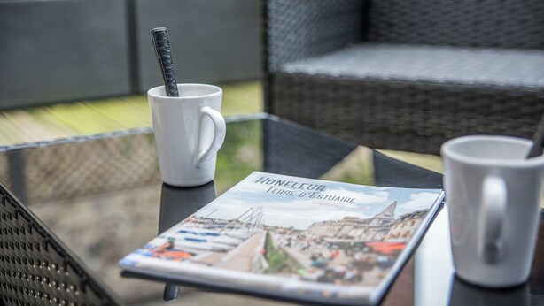 A glass table with a magazine, a white mug, and a black mug with a spoon. - La Venelle du Bassin (Honfleur)