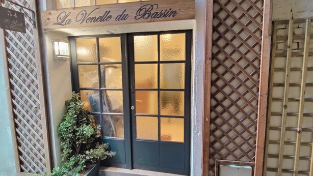 Une entrée d’immeuble avec une enseigne indiquant « La Venelle du Bassin », une porte noire dotée de panneaux en verre dépoli et une fenêtre à croisillons sur la droite.