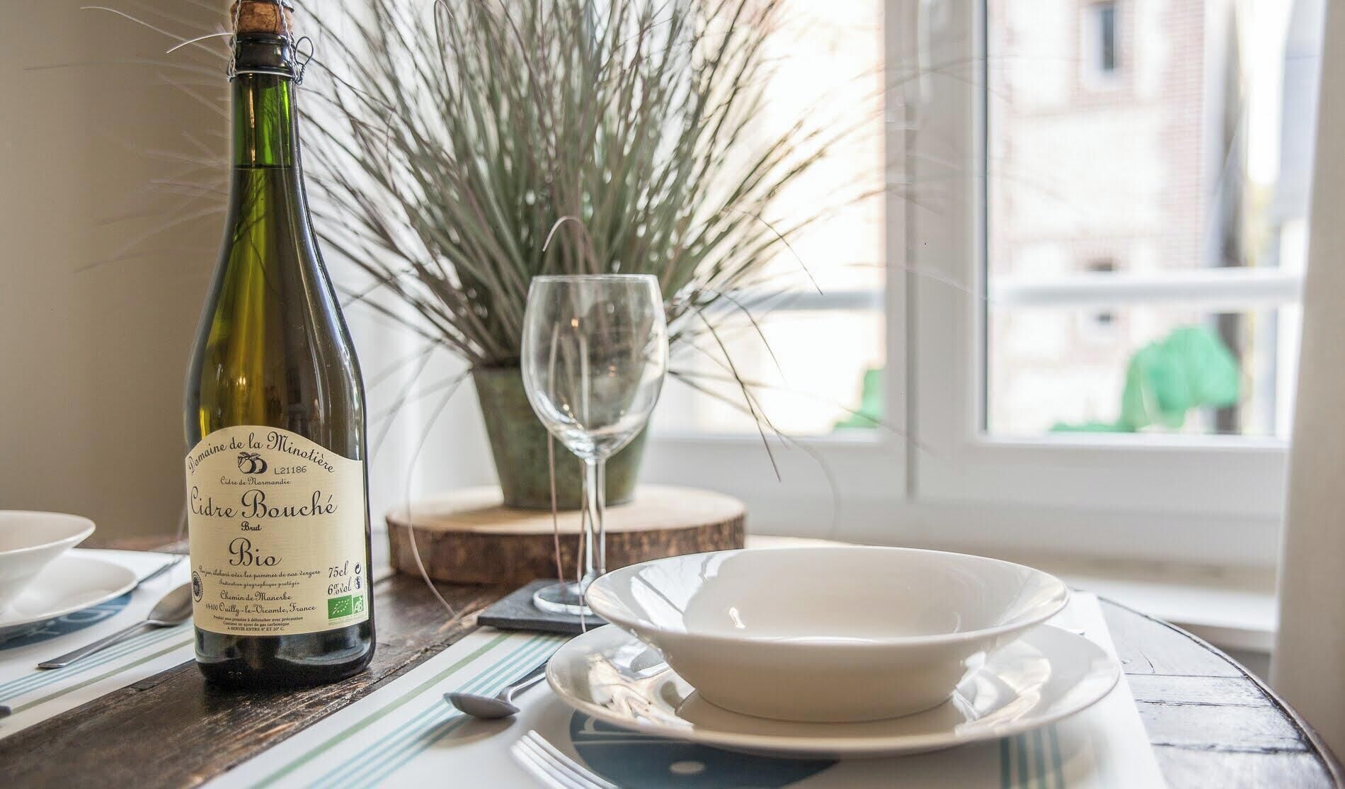 Una bottiglia di vino Domaine de la Minotière Cidre Bouché, un bicchiere e una ciotola su un tavolo con vista su un edificio attraverso una finestra.