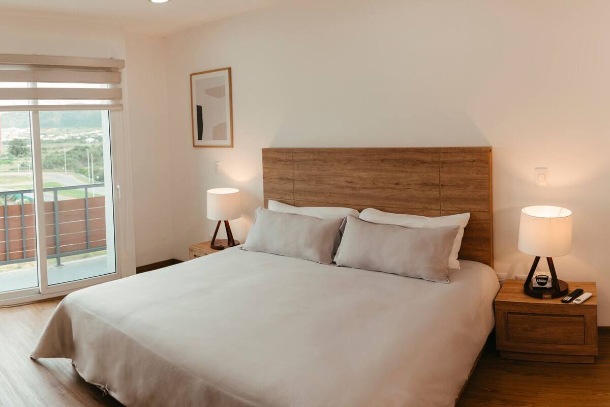 A bedroom with a large bed, two bedside tables with lamps, a window with blinds, and a framed picture on the wall.