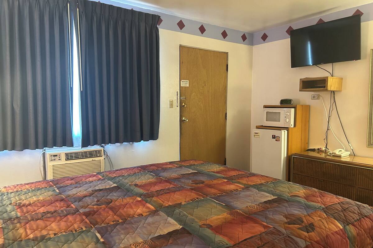 A hotel room with a bed, an air conditioner, a television, a microwave, and a wooden dresser.