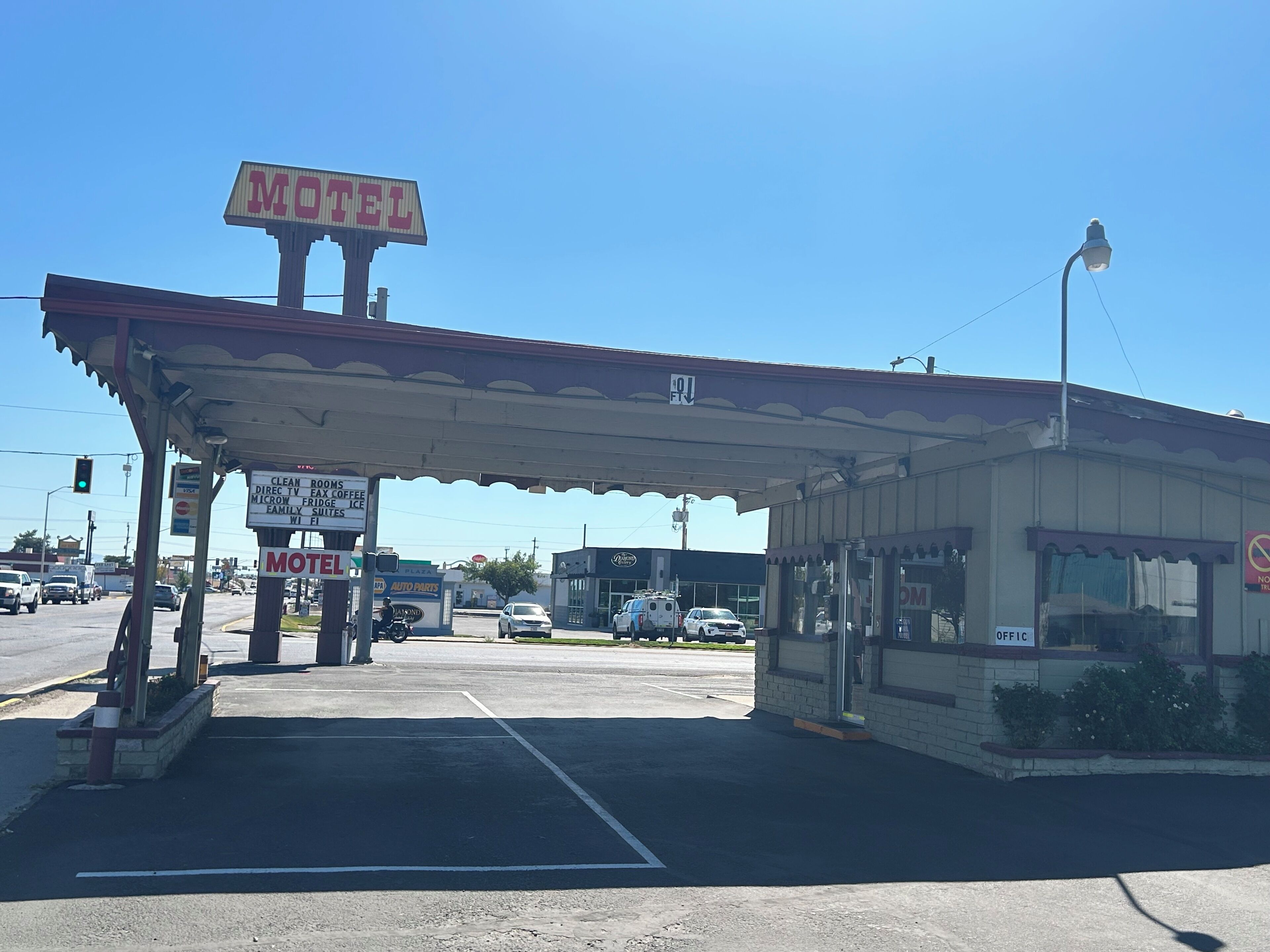 Een motel met een benzinestation, inclusief een bord met 'Motel' en voorzieningen zoals 'Schoon Kamers' en 'Magnetronbestendige IJs'.