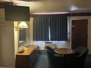 A hotel room with a television, a desk with a telephone, a chair, a round table, a window with curtains, and an air conditioning unit. - Oregon Trail Motel (Ontario)
