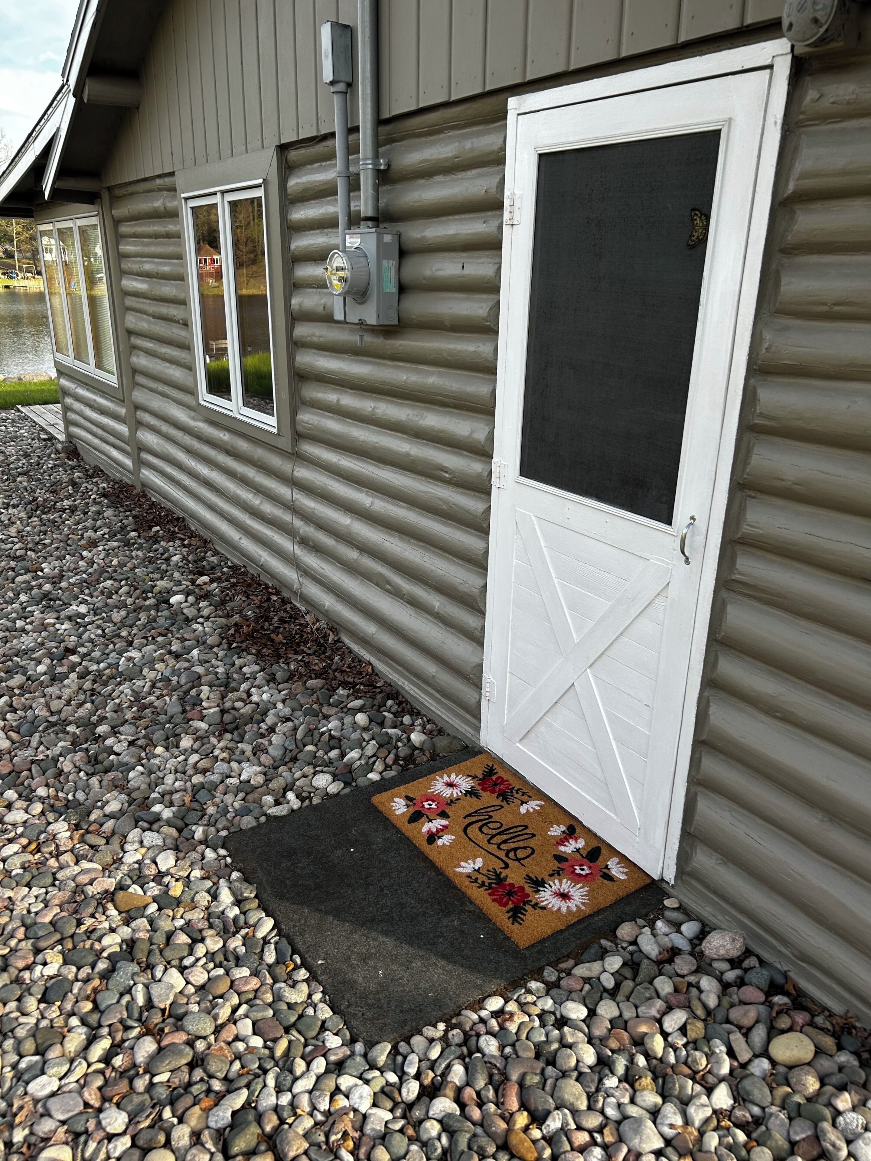 Une cabane en rondins avec une porte blanche, une fenêtre et un porche agrémenté d’un paillasson.