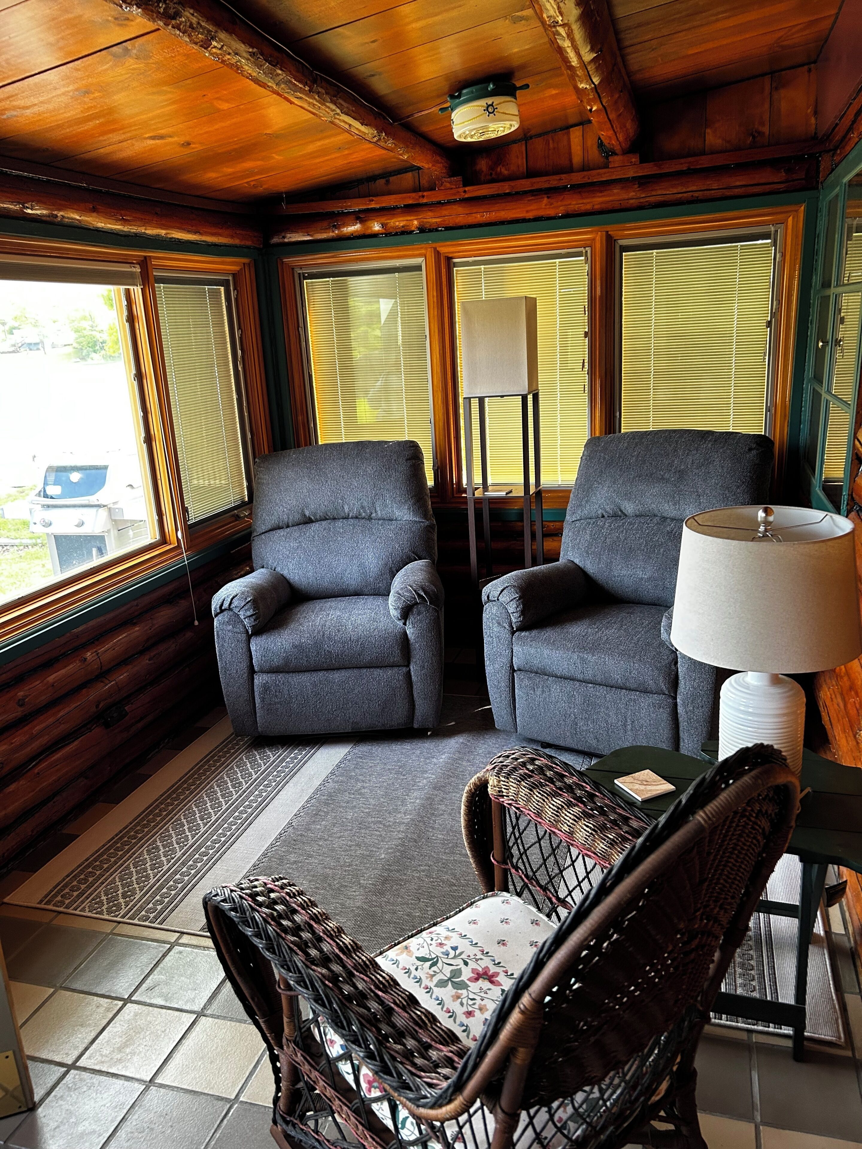 L’intérieur d’une cabane avec deux fauteuils gris, une chaise en osier, une table et une lampe.