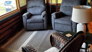 Interior de una cabaña con dos sillones grises, una silla de mimbre, una mesa y una lámpara.