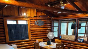 Interior de una cabaña de troncos con una mesa de comedor, sillas, un ventilador y una mesita auxiliar.