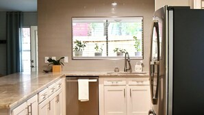 A kitchen with white cabinets, a stainless steel refrigerator, a window with plants, and a gray rug.