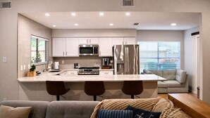 A modern kitchen with white cabinets, stainless steel appliances, a central island with bar stools, and a seating area with a sofa and window blinds.