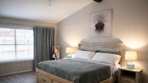 A bedroom with a bed, two bedside tables, a nightstand, a window with blinds, a bookshelf, and a painting on the wall.
