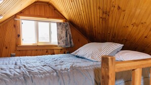 A loft bedroom with a sloped ceiling, a window with curtains, a bed with a striped pillow, and a wooden headboard. - Farmside Tiny Home (Lyman)