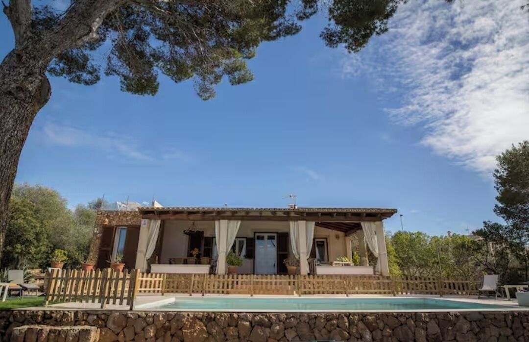 A stone-walled pool with a wooden deck, a covered patio with curtains, and a stone building with a tiled roof.