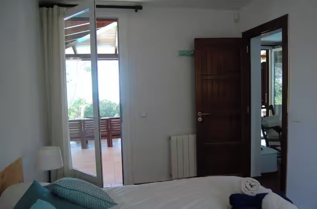 A bedroom with a bed, a window, a door, and a view of an outdoor balcony.