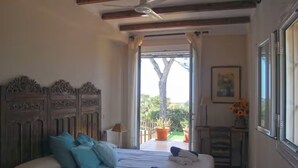 Una camera da letto con un letto, una ventola a soffitto, una finestra con tende, una pianta in vaso e un tavolino con una lampada.