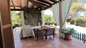 Un patio coperto con tavolo e sedie, una pianta in vaso e vista sul verde circostante.