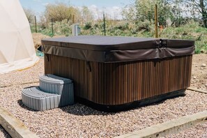 Un jacuzzi con revestimiento de madera y un escalón pequeño para acceder, rodeado de grava y una tienda de campaña al fondo.