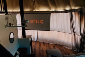 Una habitación con una pantalla grande mostrando 'NETFLIX', piso de madera, un sofá y una cortina.