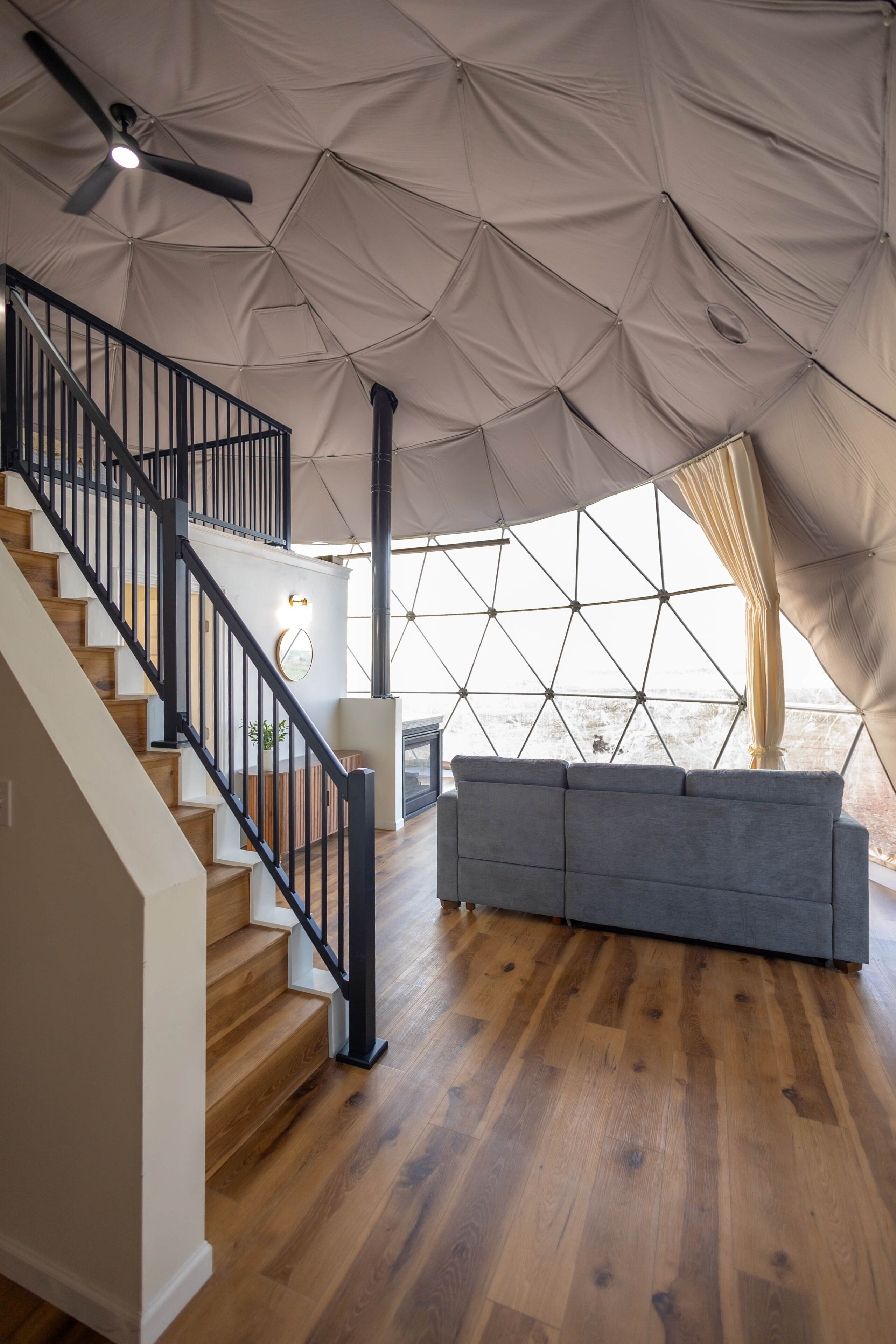 Un interior espacioso con una escalera de madera, un sofá gris y una gran estructura tipo tienda con un diseño geométrico.