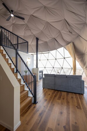 Un ambiente spazioso con una scala in legno, un divano grigio e una grande struttura simile a una tenda con motivo geometrico.