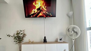 A flat-screen TV mounted on a wall displaying a fire, a white cabinet below, a small statue, a vase with plants, and a standing fan. - MARETA TORICES HOUSE (Cartagena de Indias)