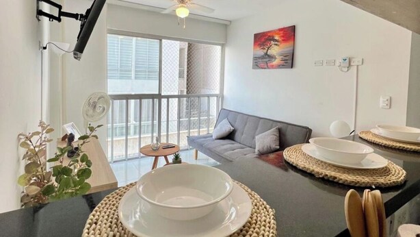 A modern living room with a grey sofa, a dining table with plates, a ceiling fan, and a large window with blinds. - MARETA TORICES HOUSE (Cartagena de Indias)