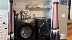 A laundry room with two front-loading washers and dryers, a metal shelf above, and a door with curtains. - Cozy 1-bedroom house with sofa sleeper, and air mattress in quaint Nashville (Nashville)