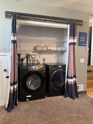 A laundry room with two front-loading washers and dryers, a metal shelf above, and a door with curtains. - Cozy 1-bedroom house with sofa sleeper, and air mattress in quaint Nashville (Nashville)