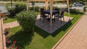 Un espacio al aire libre techado con mesa y sillas, rodeado de un jardín cuidado con césped y plantas.