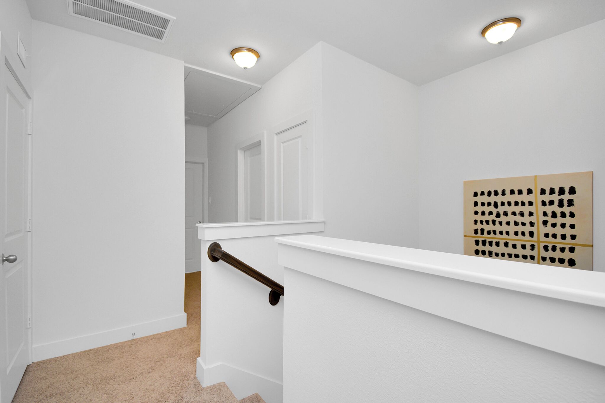 Un couloir intérieur aux murs blancs, avec un tapis beige, une porte blanche, un mur blanc, une main courante marron et un luminaire.