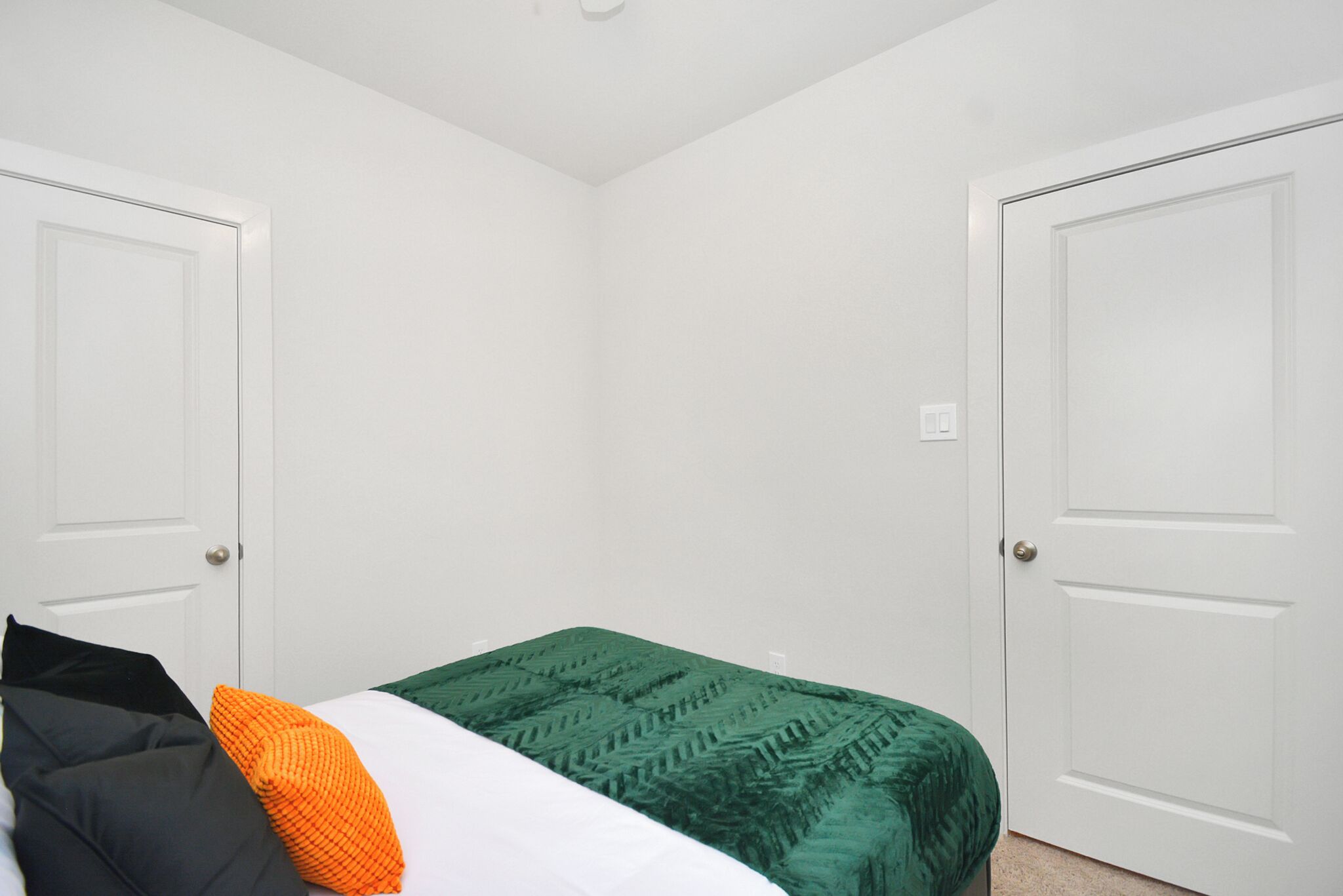 A small, white-walled room with a single bed, two doors, and a light switch on the wall.
