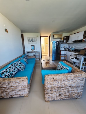 A living room with two wicker sofas, a coffee table, and a kitchen area with a refrigerator and stove. - Modern apartment with pool in Bello Horizonte | Near the beach (Santa Marta)