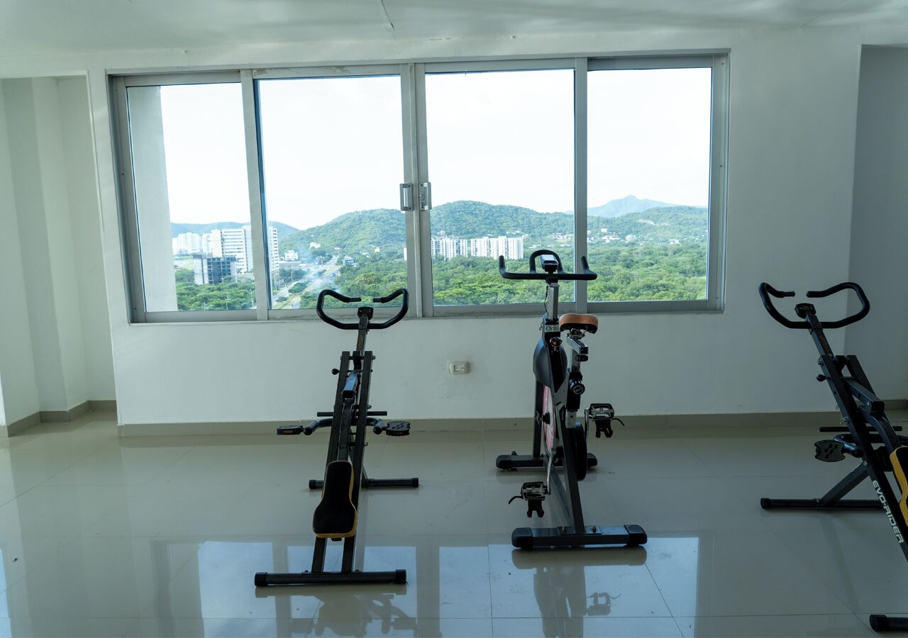 A room with three stationary bikes, large windows with a view of greenery and buildings, and a tiled floor.